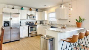 Una cocina moderna con armarios blancos, electrodomésticos de acero inoxidable, una isla central con taburetes y luminarias colgantes.