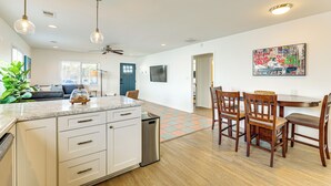 Una cocina moderna con armarios blancos, encimera de granito y lavavajillas empotrado. Hay un comedor con sillas de madera y una mesa, y un ventanal que proporciona luz natural.