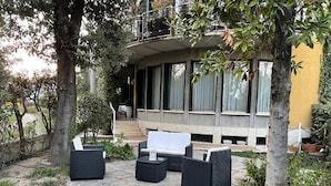 An outdoor seating area with wicker furniture, a stone pathway, and a building with large windows in the background. - Triple Room Business (Ravenna)