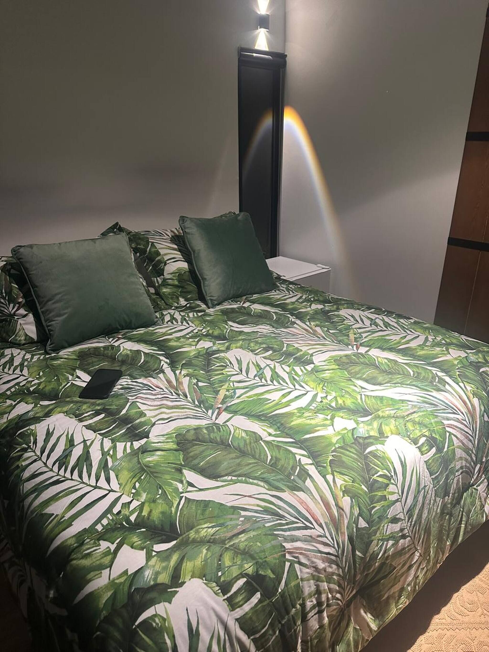 En seng med et sengetæppe med et tropisk blad-mønster, to puder, et natbord med en lampe og en vægmonteret lysarmatur.