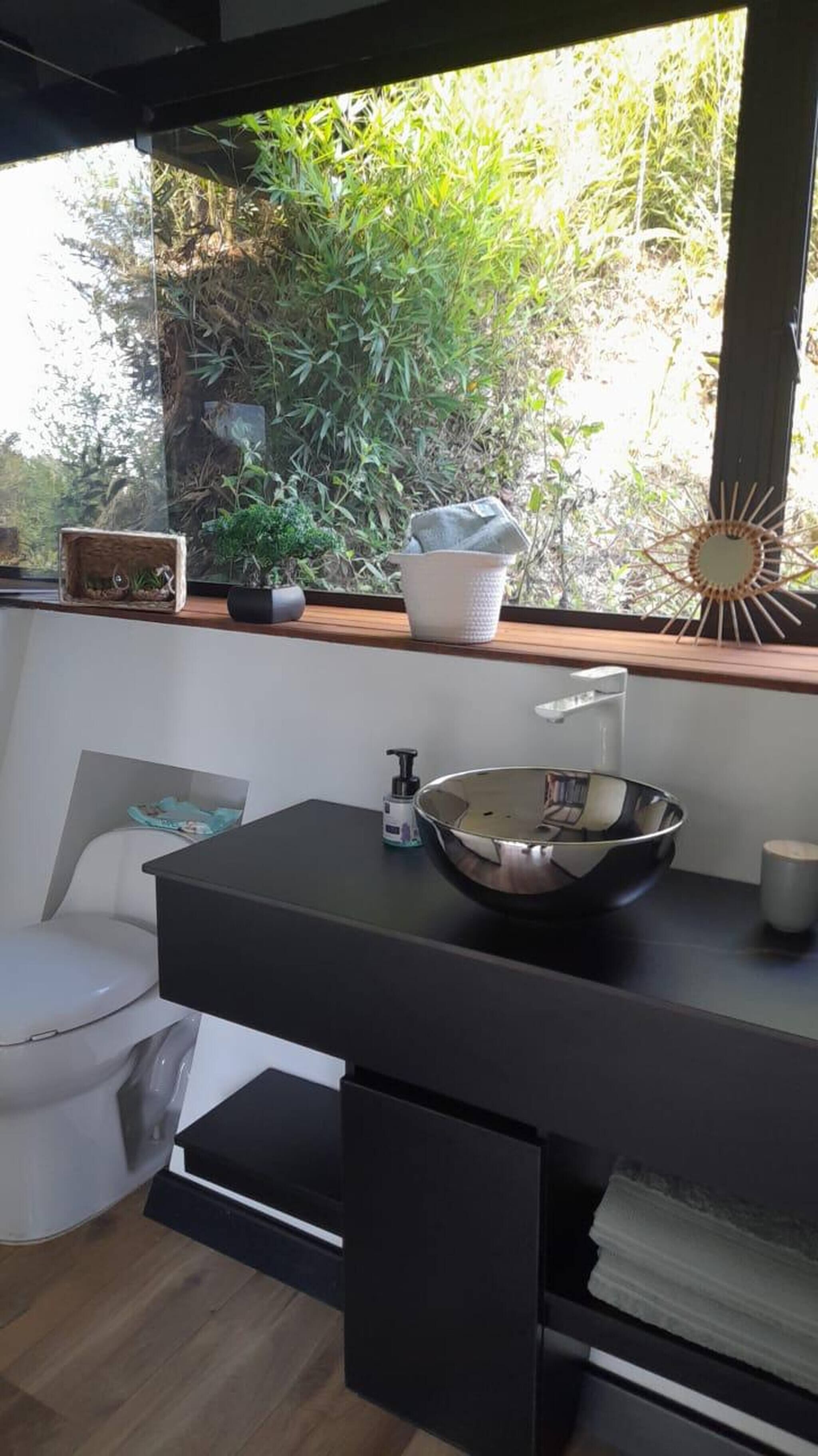 Ein modernes Badezimmer mit schwarzer Ablage, einem weißen Waschbecken und einem Fenster mit Blick auf Grünflächen.