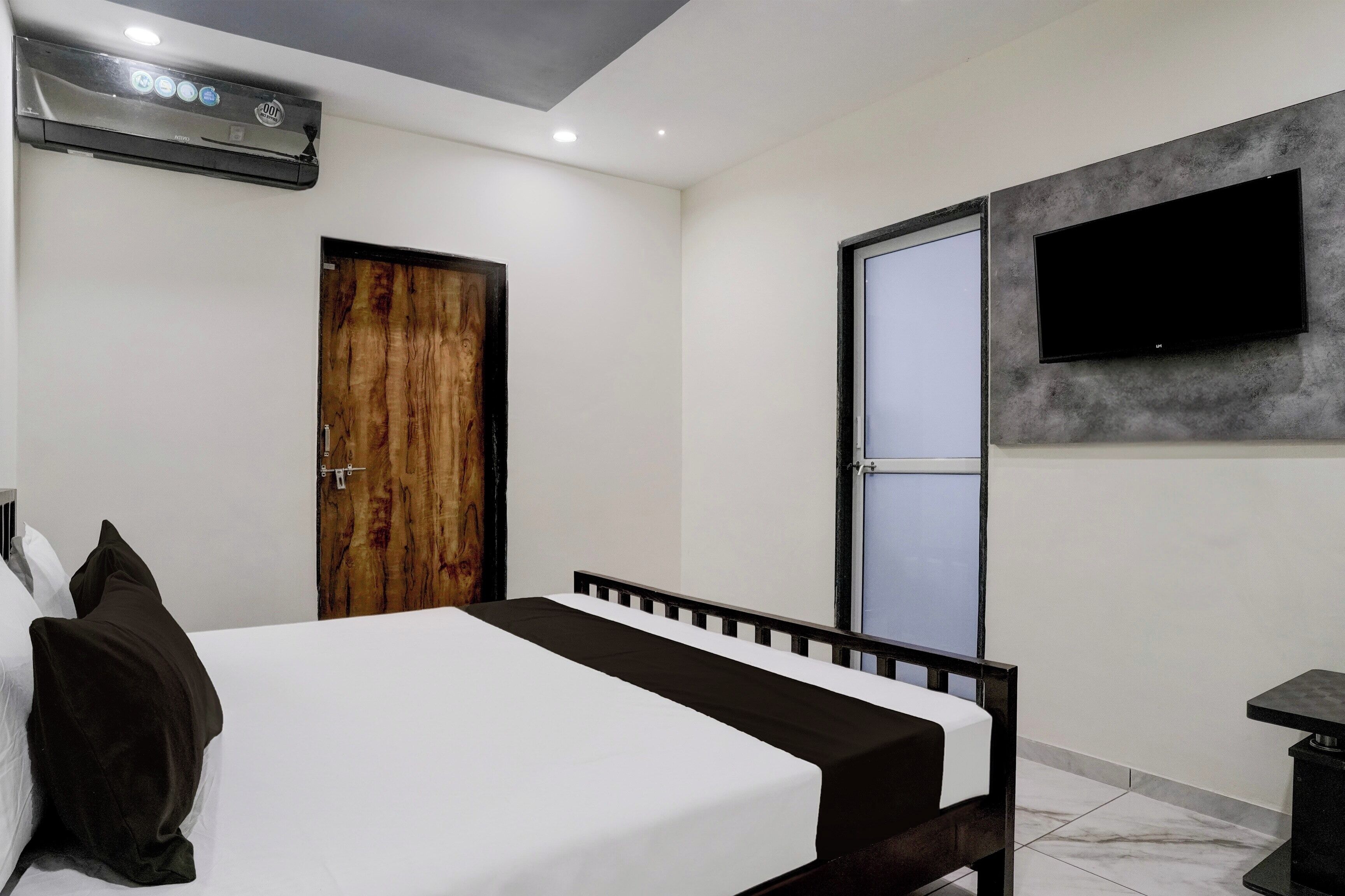 A bedroom with a bed, a wooden door, a television mounted on the wall, and an air conditioning unit.