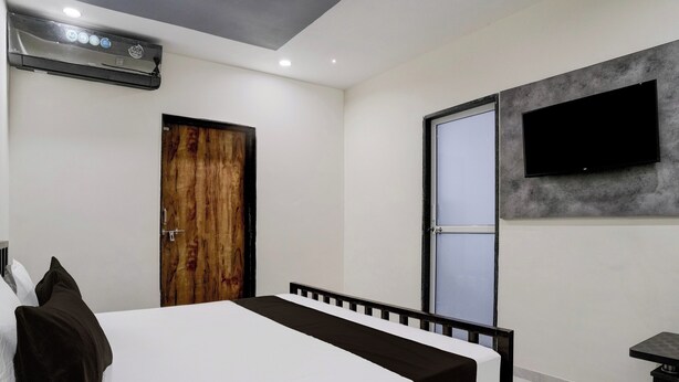 A bedroom with a bed, a wooden door, a television mounted on the wall, and an air conditioning unit. - Hotel Ambika, Vadodara (Waghoriya)
