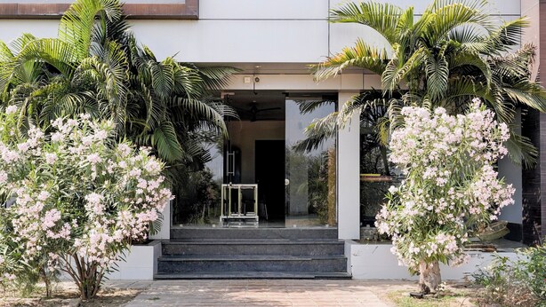 A modern building entrance with a glass door, surrounded by lush greenery and flowering plants. - Hotel Ambika, Vadodara (Waghoriya)