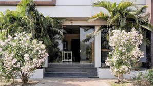 A modern building entrance with a glass door, surrounded by lush greenery and flowering plants. - Hotel Ambika, Vadodara (Waghoriya)