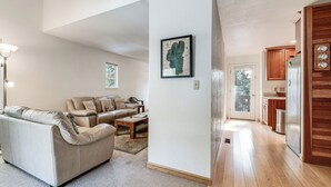 Ett rymligt vardagsrum med en beige soffa, ett soffbord och ett inramat konstverk på väggen. En trädörr leder in till ett kök med ett kylskåp och träskåp.