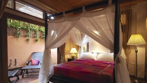 Una cama con dosel con un edredón rojo y rosa, una lámpara de noche y una mesita de noche con una lámpara. También hay un banco con cojín y una pequeña mesa con un objeto decorativo.