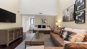Ein modernes Wohnzimmer mit Sofa, Couchtisch und Fernseher. Ein Fenster mit Jalousien und eine Wandlampe sind zu sehen.