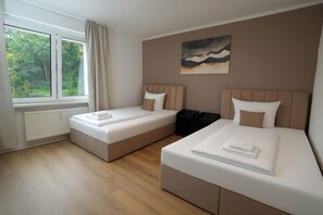 A hotel room with two beds, a large window with curtains, a radiator, and a painting on the wall.