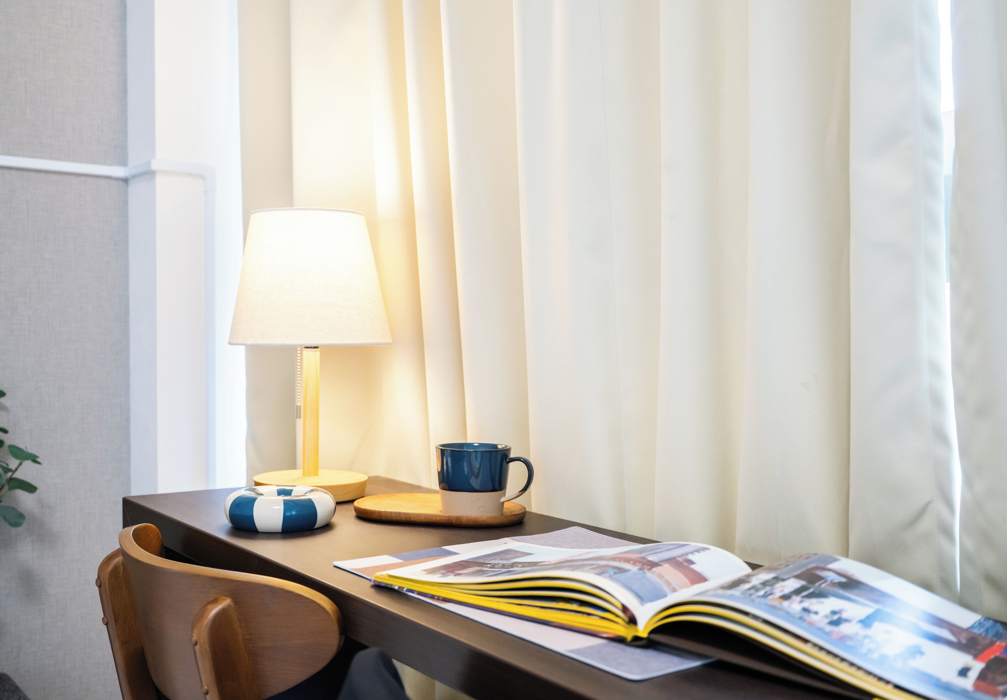 A hotel room with a wooden desk, a lamp, a blue mug, a blue and white beach ball, and an open magazine.