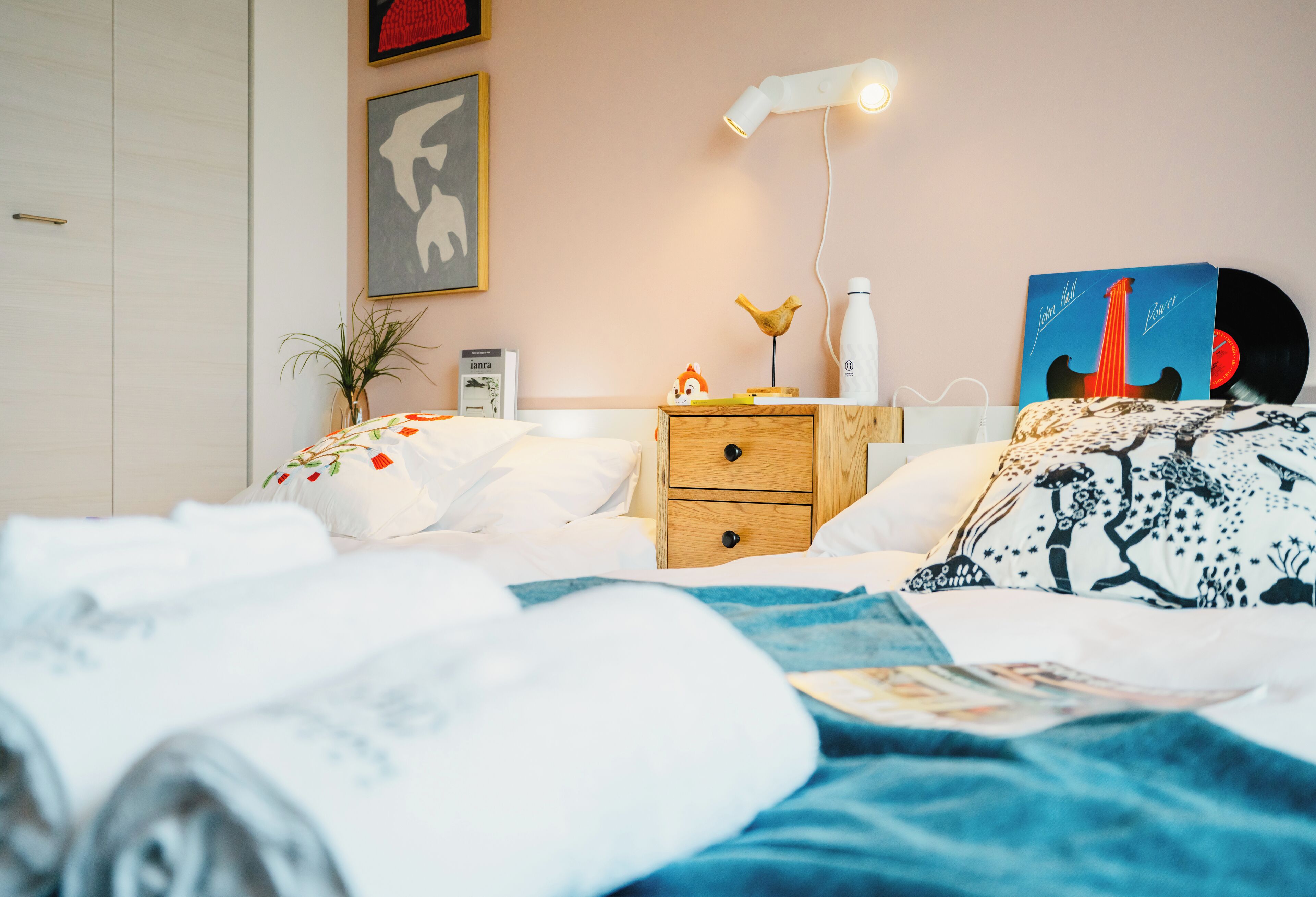 Een netjes opgemaakt bed met een deken met blauw-witte patronen, een nachtkastje met een lamp, een lijst met een afbeelding en een kleine plant.