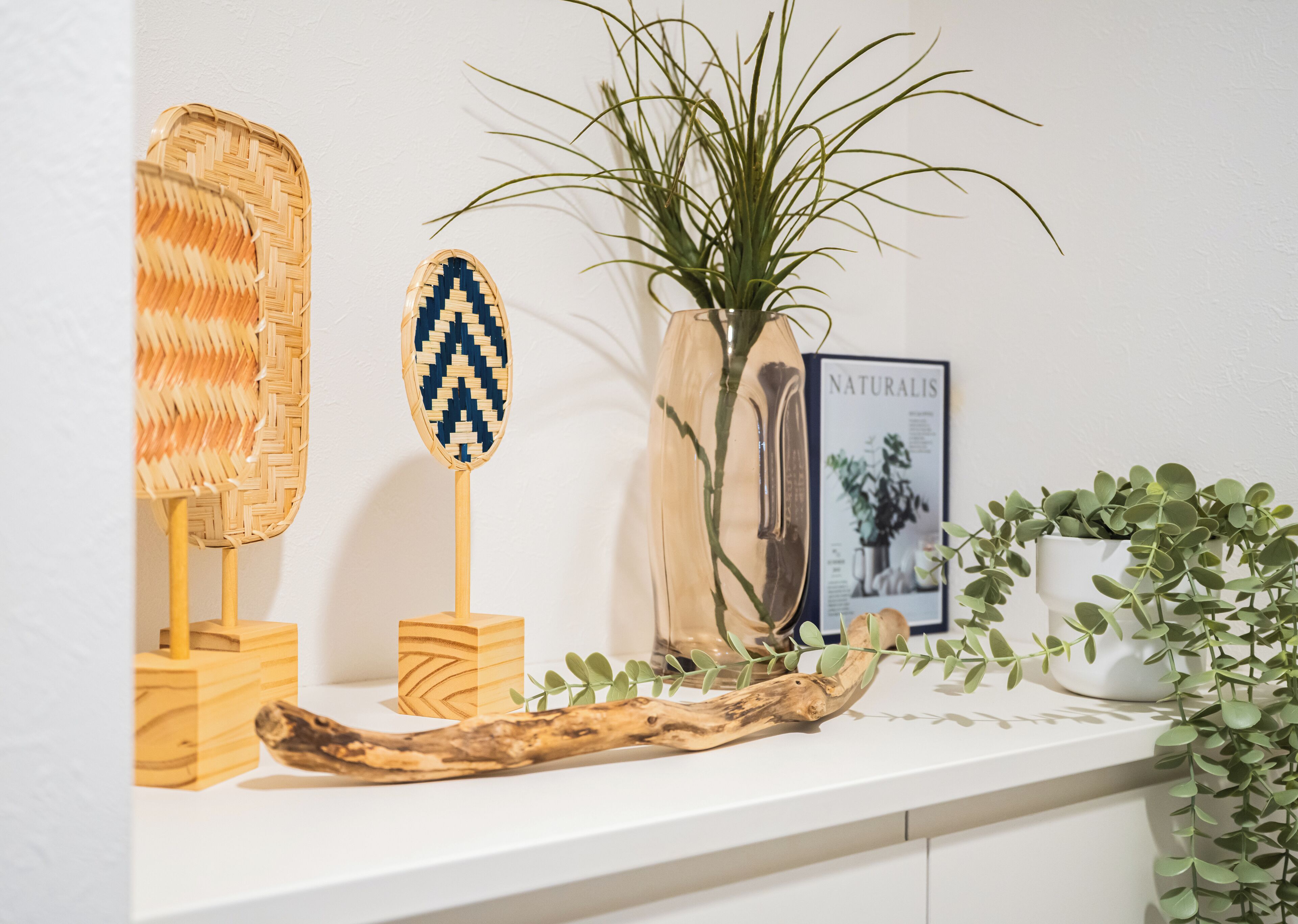 A white shelf with a woven basket, a wooden fan, a vase with a plant, a framed picture, and a small potted plant.