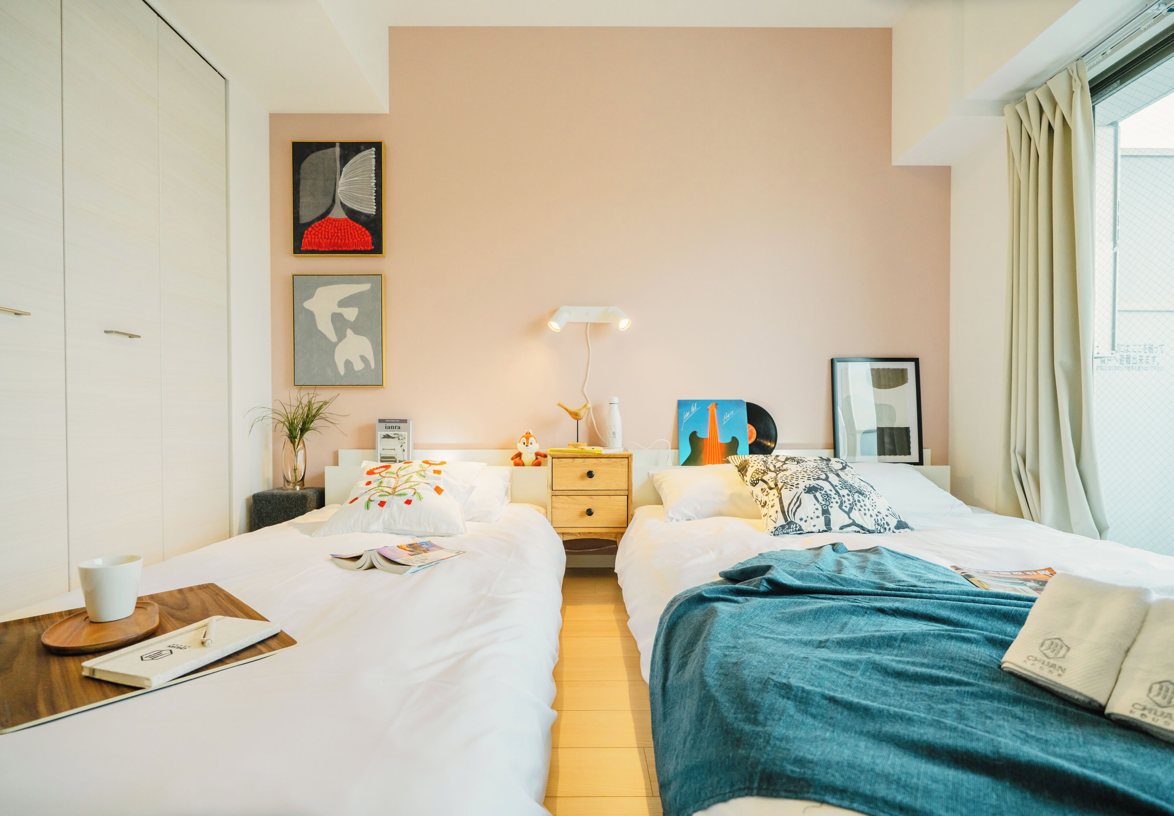 A hotel room with two beds, a bedside table with a lamp, a small plant, and artwork on the wall.
