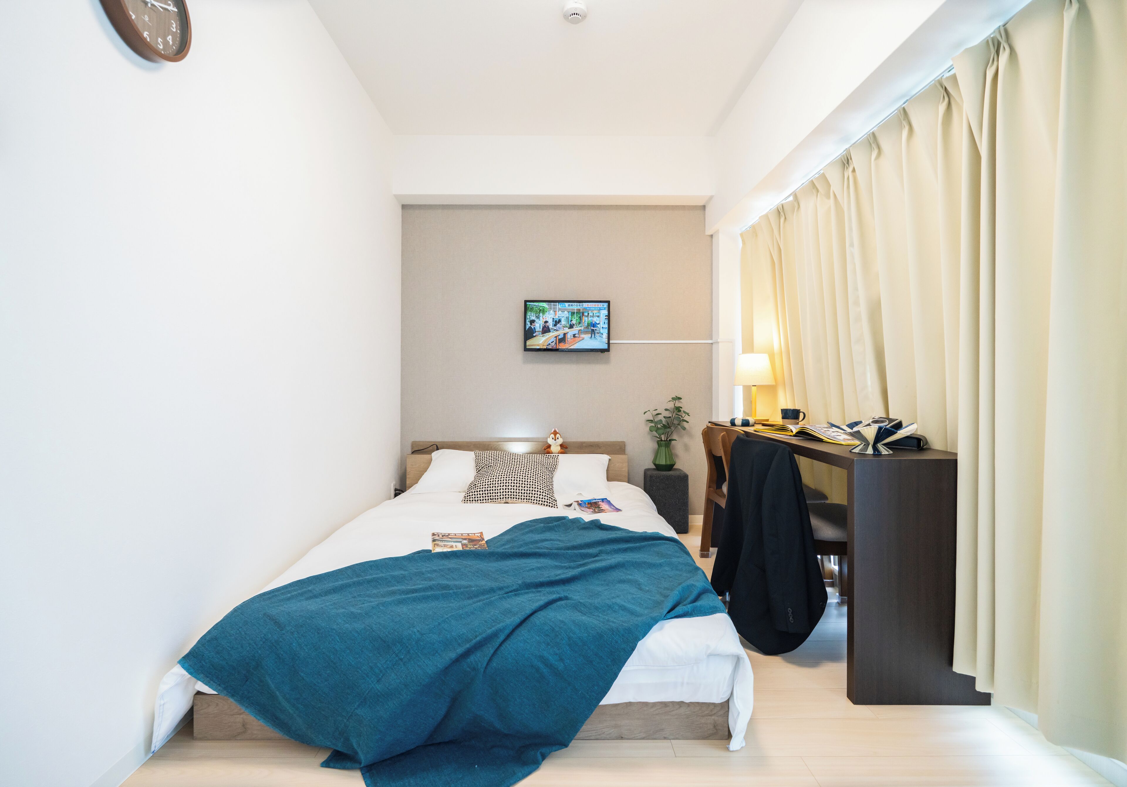 A neatly made bed with a blue blanket, a bedside table with a lamp, a TV on the wall, and a window with curtains.