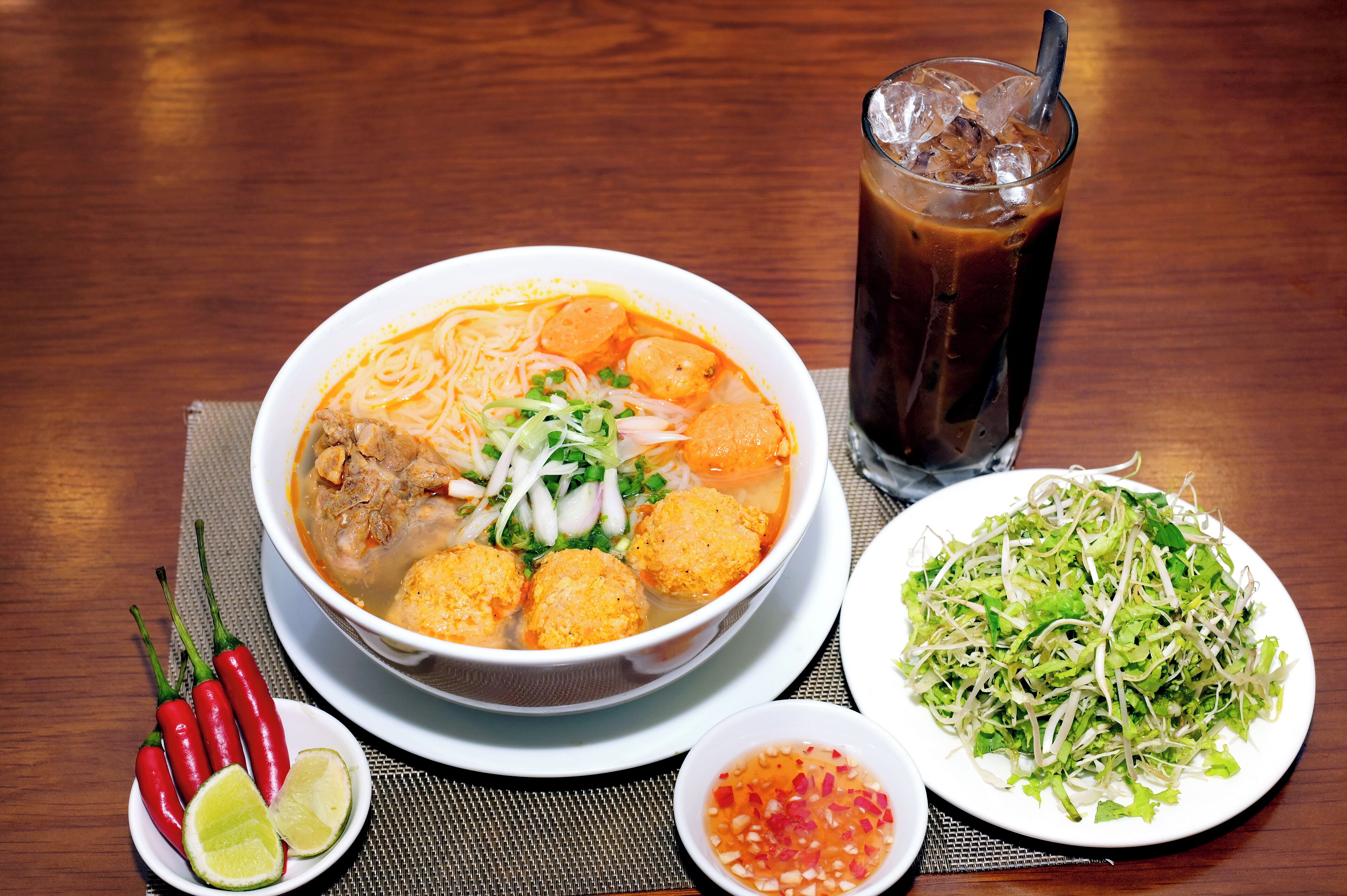 A bowl of soup with meatballs, garnished with green onions and red chili peppers, accompanied by a glass of iced beverage and a plate of fresh salad.