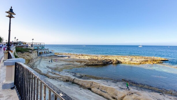 Un paseo marítimo con costa rocosa, un muelle con gente y cielo azul despejado.