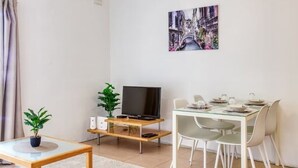 Una sala moderna con una mesa de comedor de vidrio, un televisor de pantalla plana sobre un soporte de madera, una planta en maceta y un cuadro de un paisaje urbano en la pared.