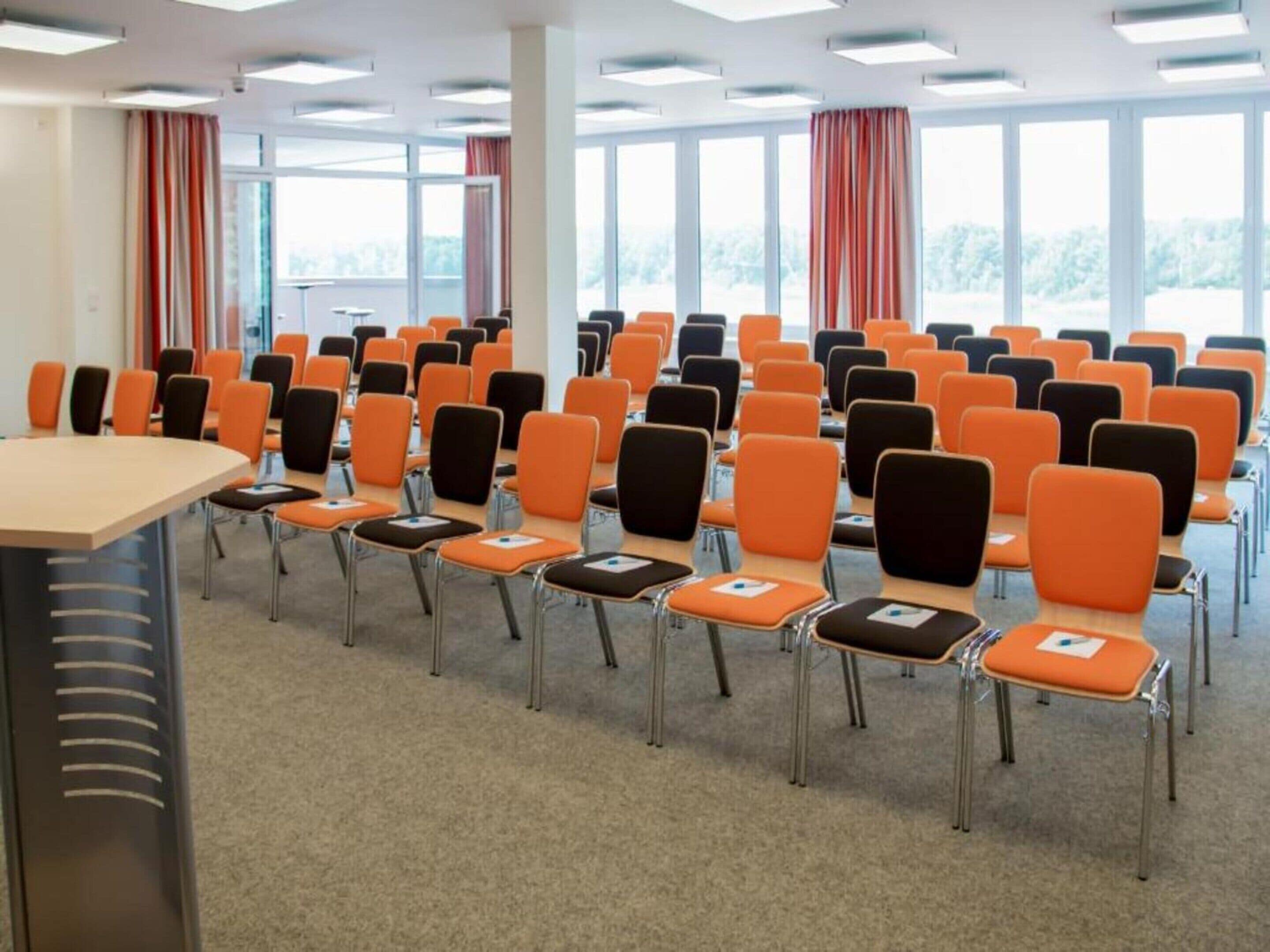 A conference room with rows of orange and black chairs, a long table, and large windows with curtains.