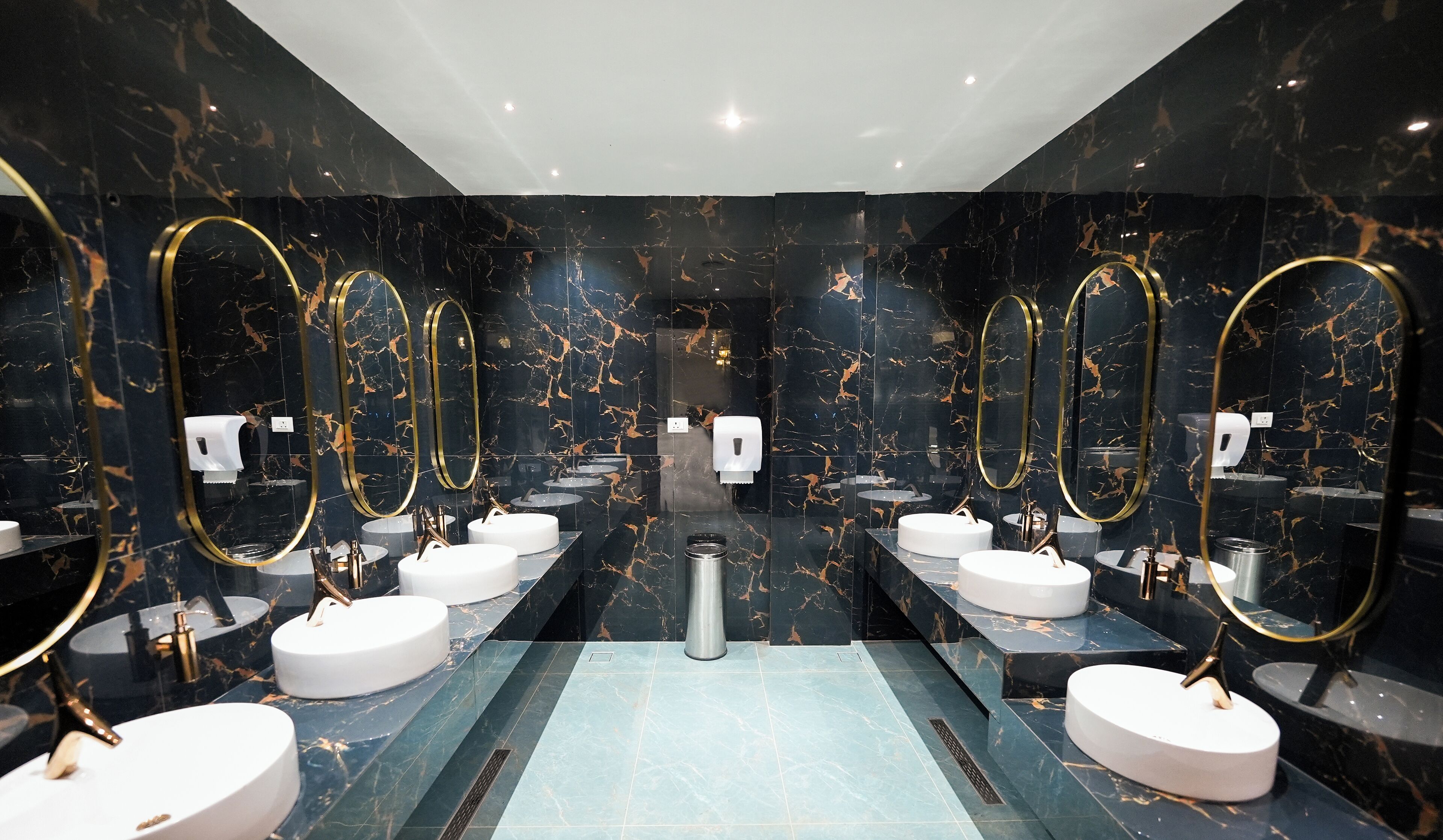 A modern bathroom with marble walls, multiple sinks, gold-framed mirrors, and a central ventilation system.