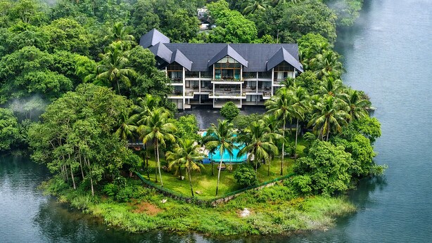 A large, multi-story resort with a pool surrounded by lush greenery and palm trees, located on an island. - Malankara palace lake view resort & spa (Thodupuzha)