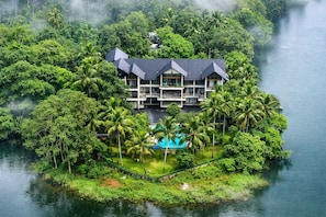 Un gran resort de varios pisos, con piscina rodeada de exuberante vegetación y palmeras, situado en una isla.