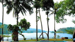 A swimming pool with a view of a lake and mountains, surrounded by palm trees and a decorative metal sculpture. - Malankara palace lake view resort & spa (Thodupuzha)