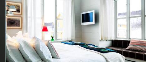 A bedroom with a bed, a TV mounted on the wall, and large windows with curtains.
