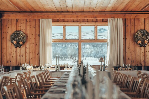 A dining area with wooden walls, a large window with a view of a snowy landscape, and a long table set for a meal. - Fjällnäs Est. 1882 (Tänndalen)