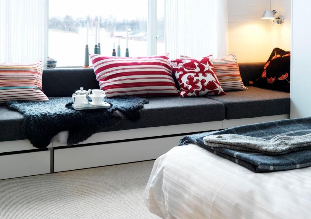 A cozy room with a sofa bed, a window with curtains, a bedside table with a lamp, and a view of the outdoors.