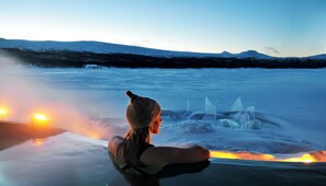 A person sitting by a fire in a snowy landscape with mountains in the background. - Fjällnäs Est. 1882 (Tänndalen)