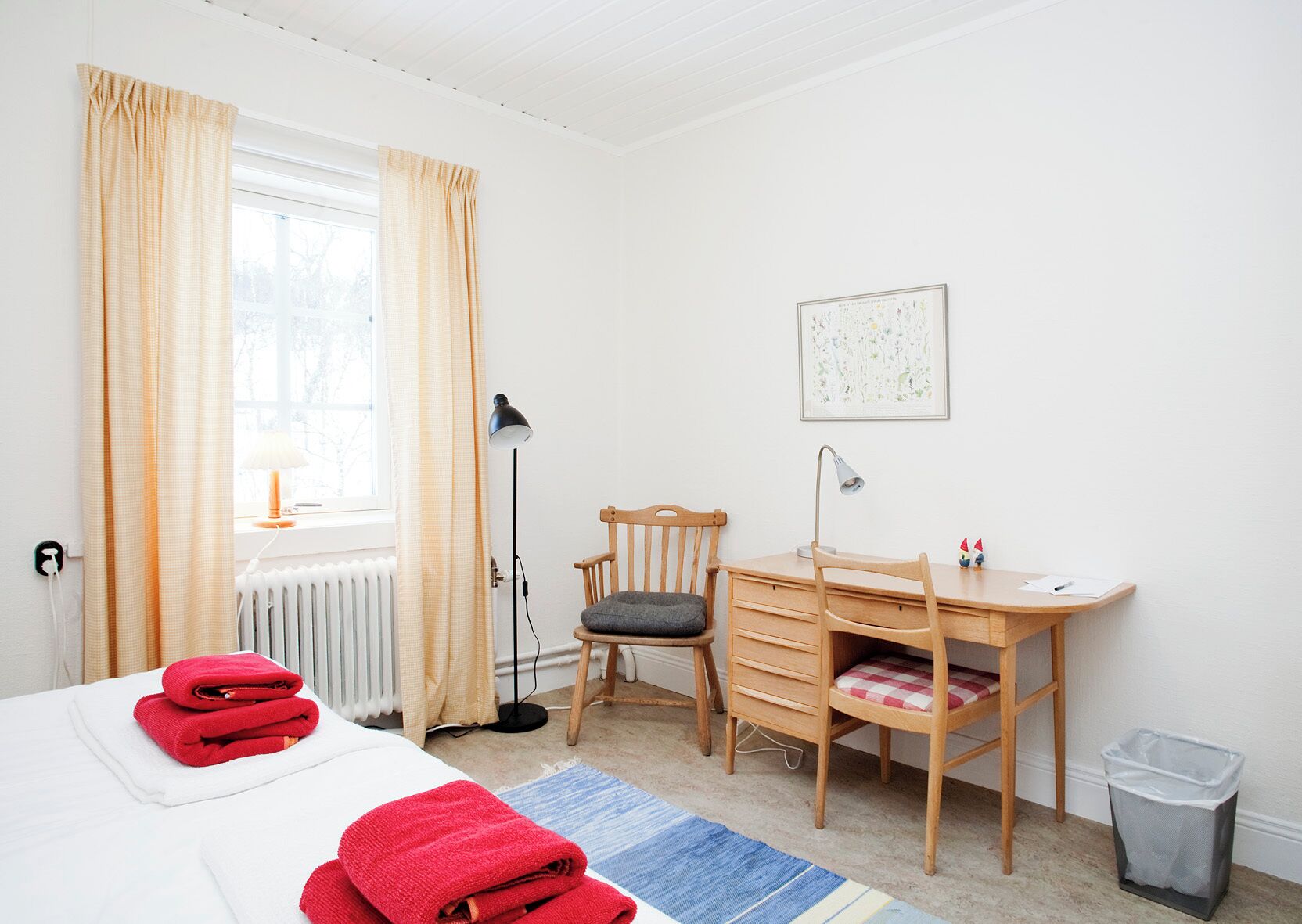 Quarto de hotel com cama, escrivaninha, cadeira, radiador, luminária, um quadro na parede e lixeira.