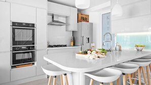 Une cuisine moderne avec des meubles blancs, un îlot central doté de tabourets, un four encastré et un luminaire suspendu.
