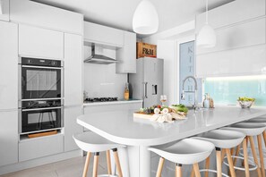 Une cuisine moderne avec des meubles blancs, un îlot central doté de tabourets, un four encastré et un luminaire suspendu.