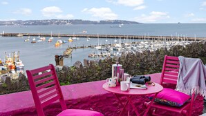 Une table recouverte d’une nappe rose, deux chaises roses, et une vue sur un port avec des bateaux et une jetée.