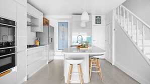 Une cuisine moderne avec des meubles blancs, un îlot central doté de tabourets, un four encastré et un luminaire suspendu.