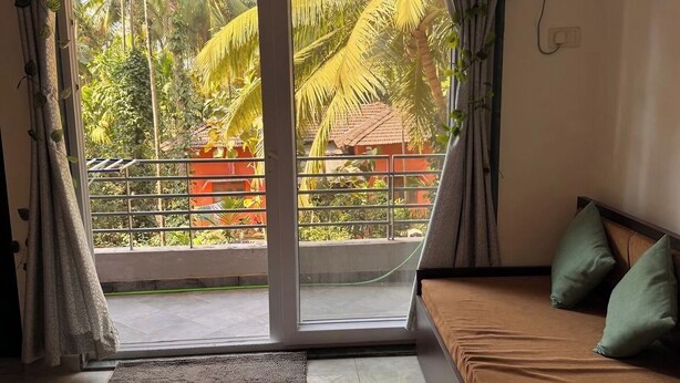 Una habitación con puerta de vidrio corredera que da a un balcón, un sofá con cojines, una alfombra pequeña y un equipo de aire acondicionado en la pared.