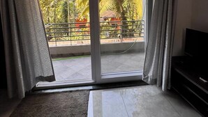 Una habitación con puerta de vidrio corrediza que da a un balcón con vistas a palmeras y un edificio rojo. Hay un ventilador de techo, un televisor y una alfombra estampada en el suelo.