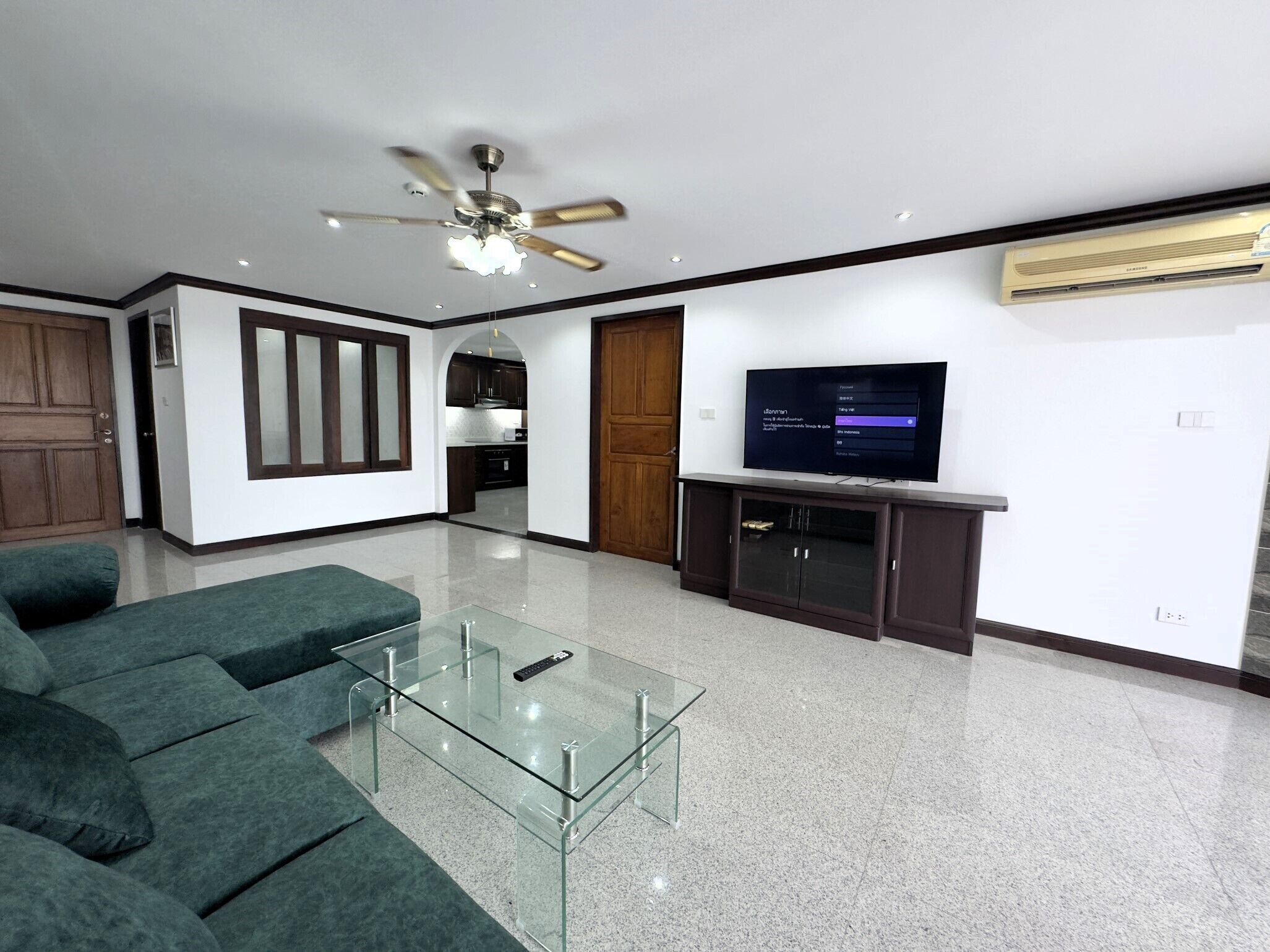 A modern living room with a glass coffee table, a sofa, a ceiling fan, a TV on a stand, and a kitchen in the background.