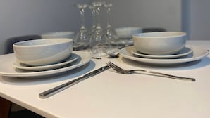 A table set with white plates, bowls, and cutlery, with wine glasses in the background. - Modern 2-Bed, Swindon Old Town (Swindon)