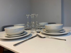 A table set with white plates, bowls, and cutlery, with wine glasses in the background. - Modern 2-Bed, Swindon Old Town (Swindon)