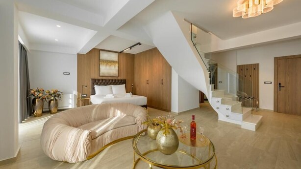 Une chambre d’hôtel moderne avec un canapé incurvé, une table basse en verre, un lit doté d’une tête de lit en bois et un escalier avec des rampes en verre.