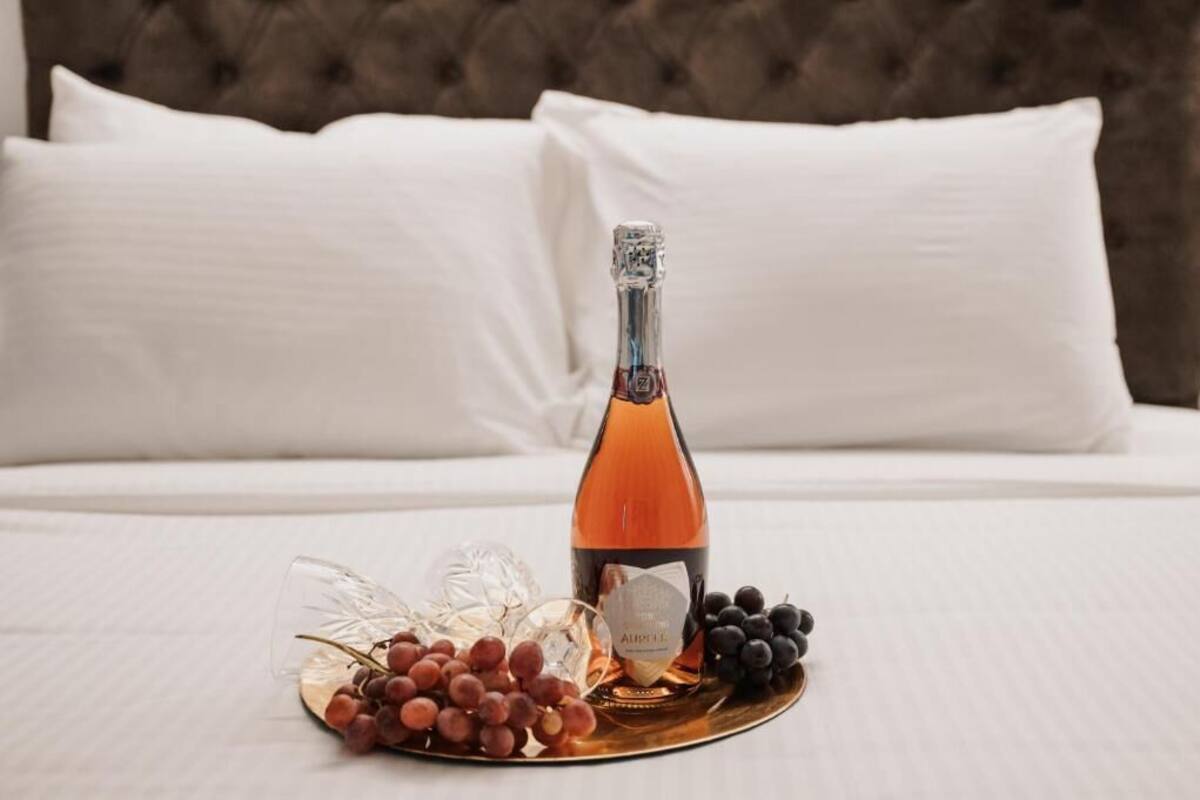 Een bed met witte lakens en een gestuufd hoofdeinde, een fles wijn en een fruitschaal met druiven en bessen.