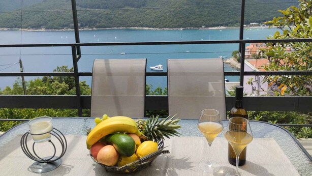 Ein Tisch mit Obstschale, zwei Weingläsern und einer Flasche auf einem Balkon mit Blick auf die Küste.