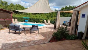 Een terras met een zwembad, buitenmeubels en een overdekt gedeelte.