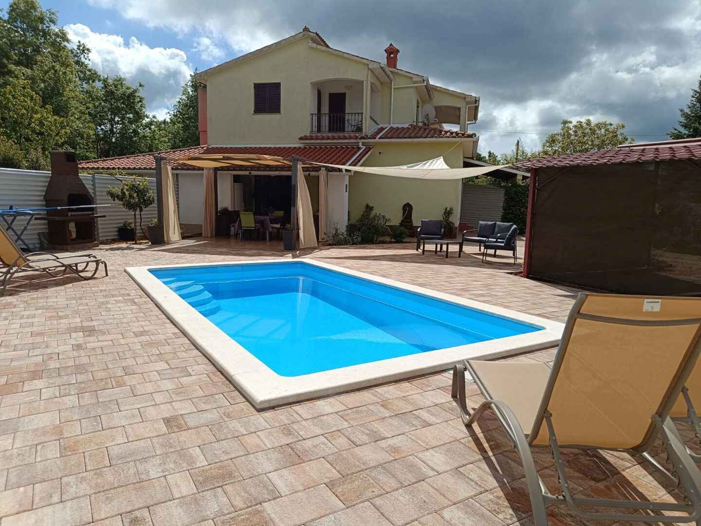 Une piscine rectangulaire avec une terrasse carrelée, un espace barbecue et du mobilier d’extérieur.