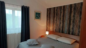 Een slaapkamer met een bed, twee kussens, een nachtkastje met een lamp, een raam met gordijnen en een houten wandpaneel.