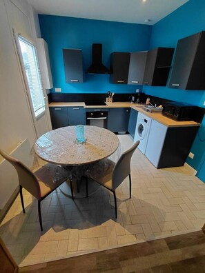A compact kitchen with blue walls, a round table with chairs, a washing machine, and a coffee machine. - Saint Malo Beautiful single-storey apartment 300 m from the station near the intramural sillon beach (Saint-Malo)