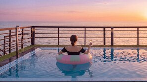 Iemand zit op een zwemband in een zwembad, met uitzicht op zee bij zonsondergang.