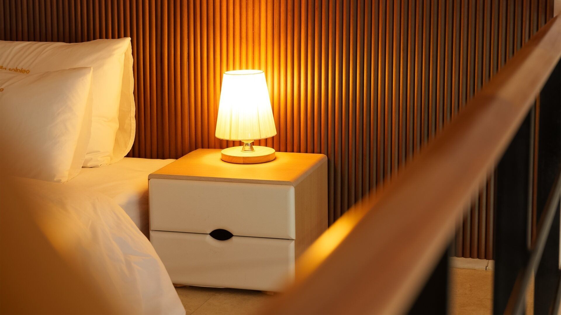 A neatly made bed with white linens, a bedside lamp, and a nightstand with a drawer.