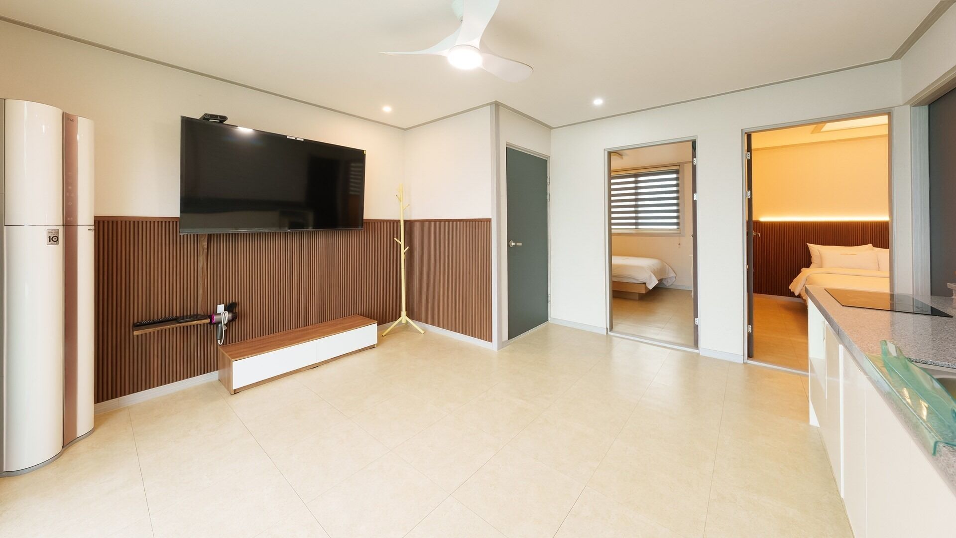 A modern hotel room with a flat-screen TV, a wooden panel wall, a built-in bench, a bed, and a bathroom visible through an open door.